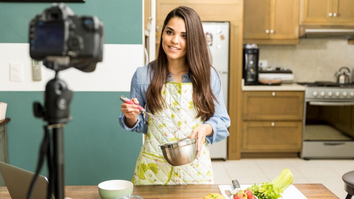 Woman in apron whisks ingredients in a bowl while filming in a kitchen.