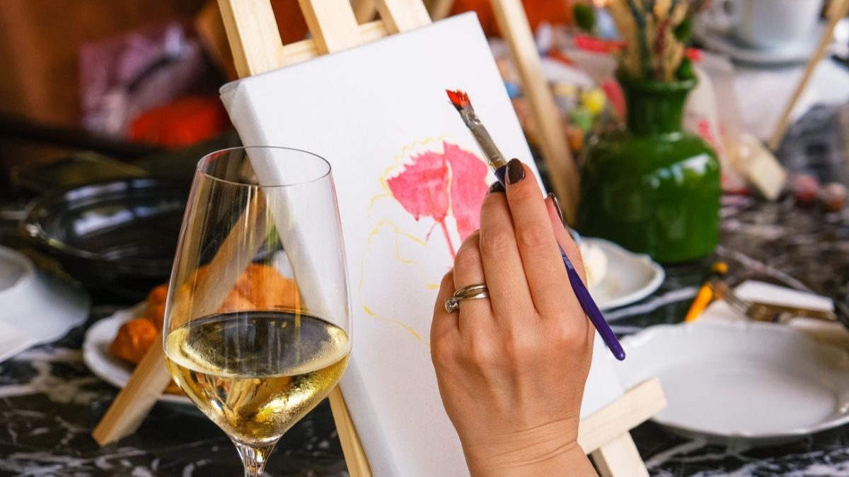 A hand paints a flower on a canvas with a glass of white wine nearby.