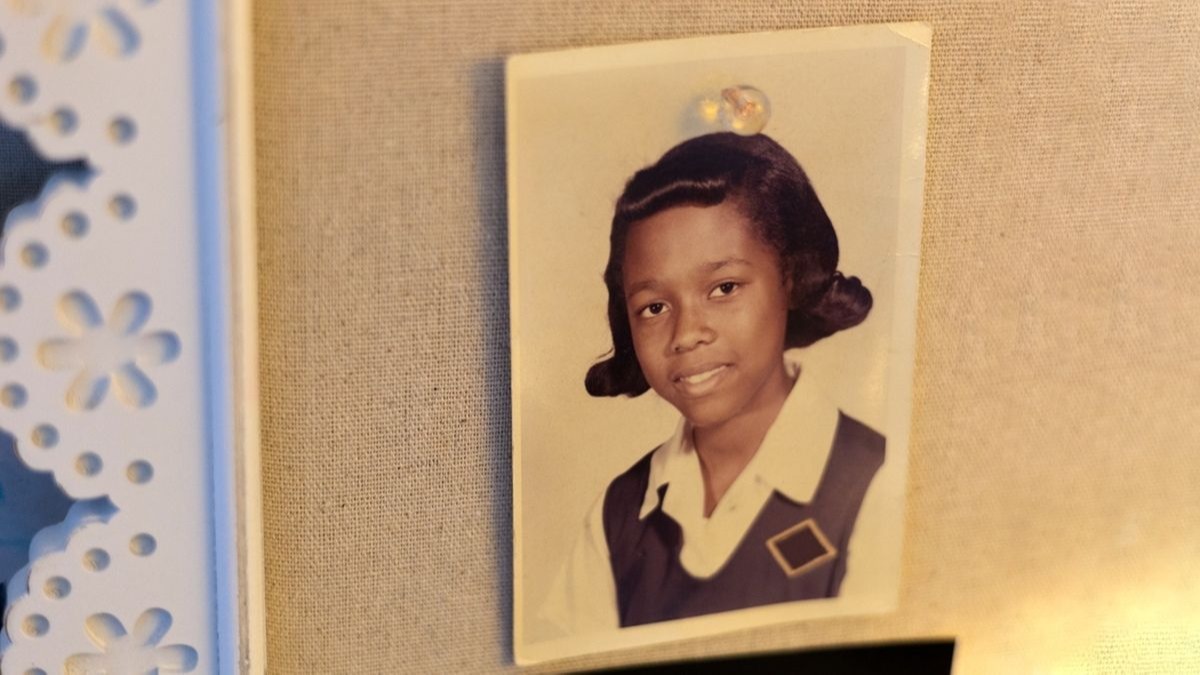 A young Black girl smiles in a school photo, wearing a white shirt and dark vest with a diamond emblem.