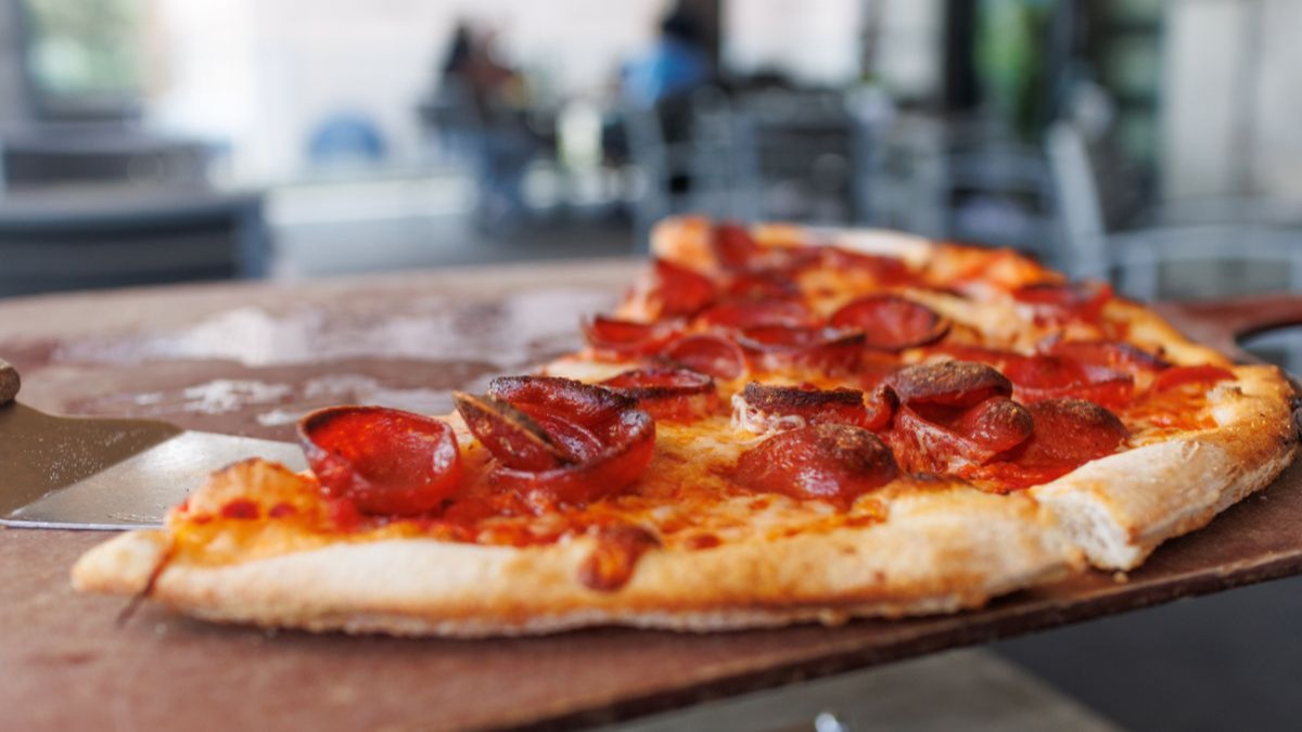 A slice of pepperoni pizza on a wooden peel, with a metal spatula on the left.