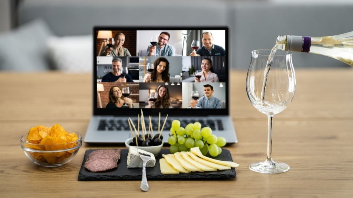 A laptop displays a video call with nine people holding wine glasses. White wine is poured into a glass in the foreground.